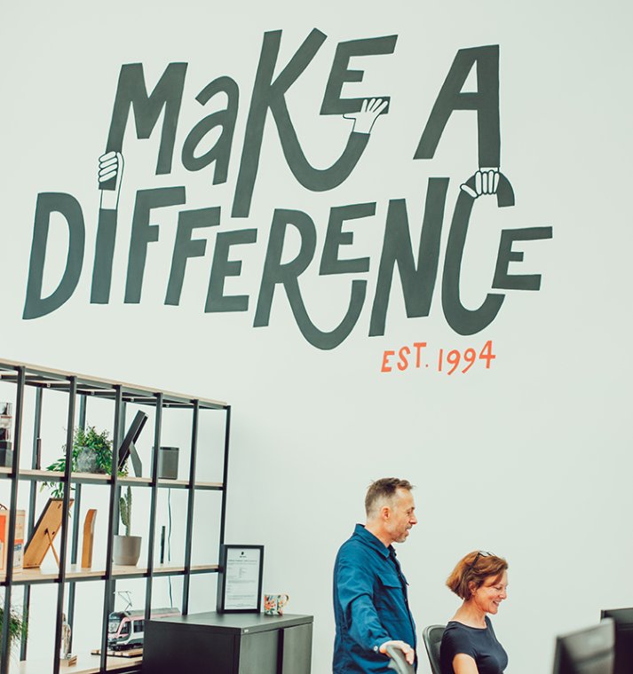 Mural on the wall shows "Make a difference. Est. 1994." In the foreground, members of the IE team at work in the studio. 